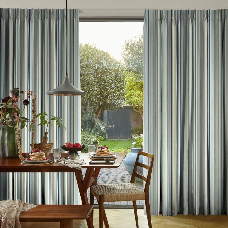 Striped pleated curtains, blue-gray and cream stripes, hang either side of a tall glass door, partially open, filtering morning light onto a dining table set for breakfast; sunlit garden beyond.