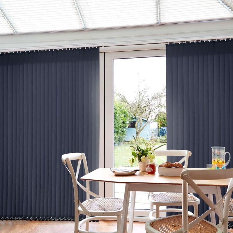 Navy vertical blinds, floor-to-ceiling and slatted, mostly closed but parted centrally to expose a glass patio door, filtering daylight into a dining area with wooden table, chairs, and garden view.