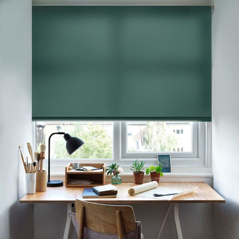 Dark green roller blind, lowered across the window with a bottom gap. Blocking and diffusing daylight. In a bright study: wooden desk with lamp, plants, art tools, suburban view.