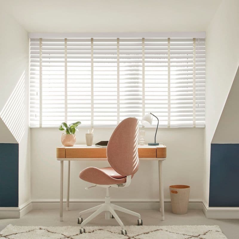 White horizontal slatted blinds with beige ladder tapes covering a dormer window — closed, filtering bright daylight — above a compact home office: wooden desk, pink swivel chair, lamp, plant, neutral walls.