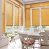 Honey‑orange horizontal slatted blinds cover tall bay windows and French doors, mostly lowered and filtering sunlight; bright dining area with white table, chairs with floral cushions and garden view.