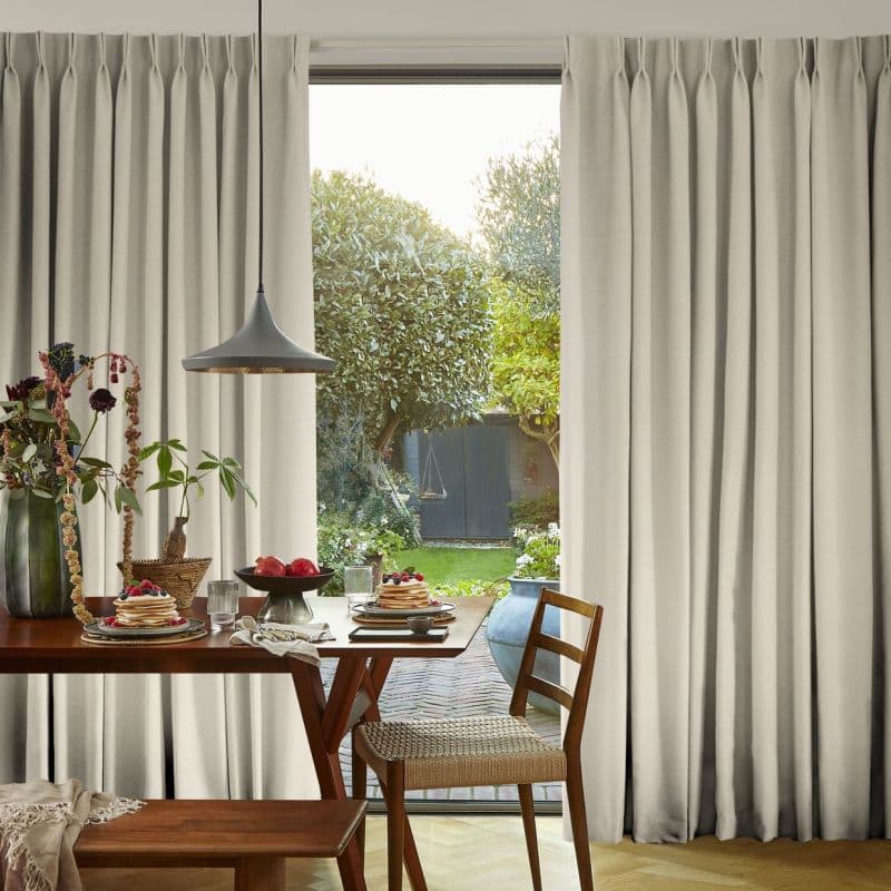 Full-length beige pleated curtains, hung floor-to-ceiling and parted at the center, framing sliding glass door; sunlight filters through onto wooden dining table set for breakfast with pendant lamp and garden view.