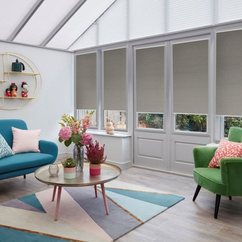Beige fabric roller blinds over tall windows and doors, lowered mid-height filtering daylight; in a bright conservatory living room with colorful chairs, a coffee table, flowers and a garden view.