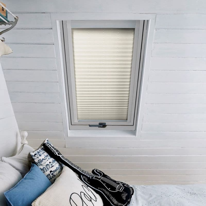 A beige pleated cellular blind fills a skylight, fully lowered and diffusing light, set in white-painted slanted wooden ceiling above a bed with blue cushions and a black guitar pillow.