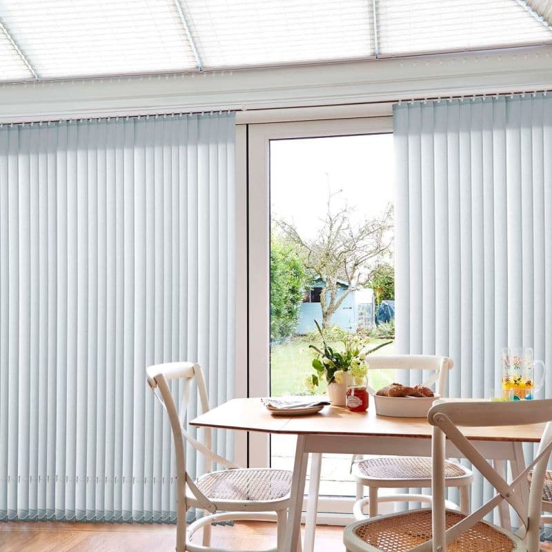Pale grey vertical blinds over a sliding glass door, parted centrally; they filter bright daylight while revealing a garden and small dining area with light wood table and cane chairs.