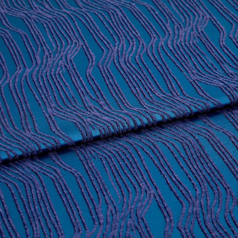 Teal fabric with raised purple wavy stripes; folded across the center, the textile lies flat with a central overlap; close-up studio view with soft, even lighting.
