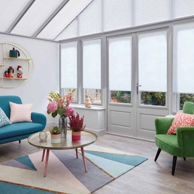 White fabric roller blinds across tall conservatory windows and door, fully lowered and diffusing soft daylight; living-room setting with teal sofa, green armchair, floral centerpiece, and pastel geometric rug.