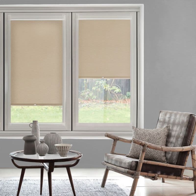 Beige roller blinds, mounted inside twin window frames, partially lowered and filtering daylight; in a softly lit living room with a rattan armchair, patterned cushion, and round coffee table.