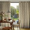 Floor-length pleated floral curtains in pale yellow and grey, partially drawn to reveal a patio door, filtering soft daylight over a dining table set for breakfast with a garden view.