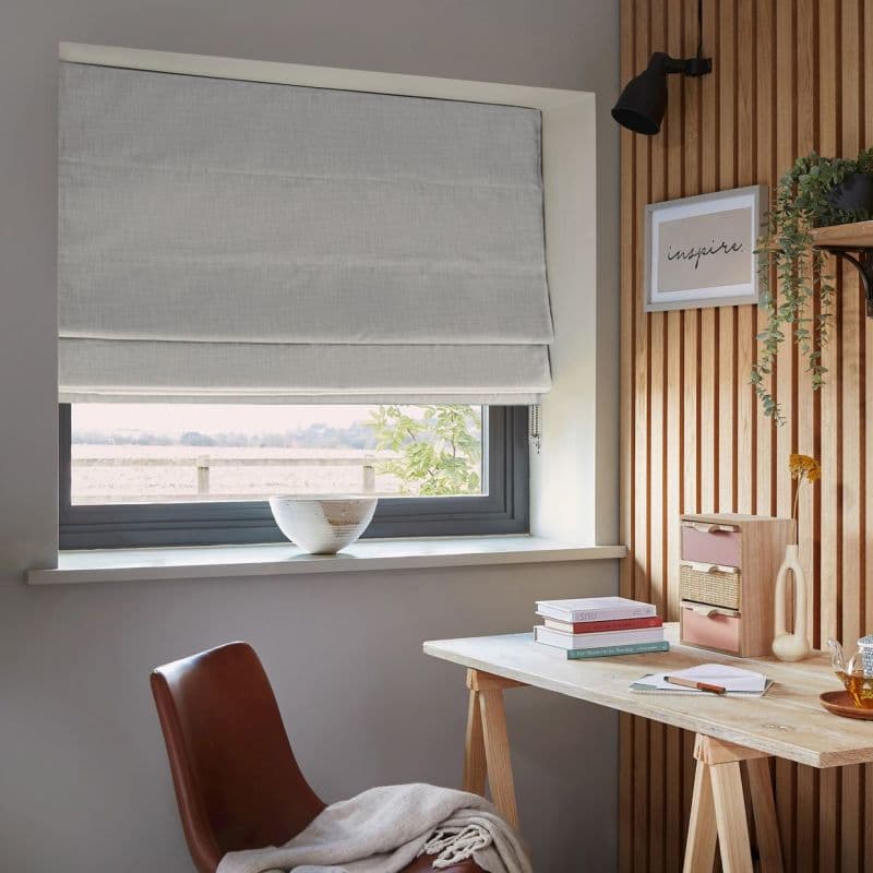 Light grey Roman blind, lowered and softly folded, filtering daylight through a grey-framed window; home office corner with wooden slat wall, desk, books, bowl on sill, leather chair and trailing plant. Framed print reads: "inspire."