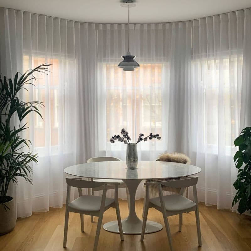 Sheer white floor-to-ceiling curtains on a curved bay of three windows, gently diffusing daylight; round marble-topped dining table with chairs and vase on a warm wooden floor.