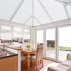 Object: white pleated blinds covering windows, French doors and pitched conservatory roof. Action: lowered, filtering and diffusing bright daylight. Context: airy kitchen-dining conservatory with wooden table and patio garden view.