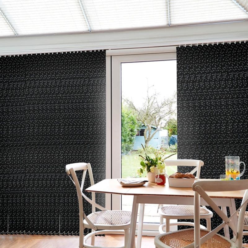 Dark patterned vertical blinds at a patio door, drawn closed at the sides and parted centrally, framing a sunlit garden; dining table with white chairs, flowers, and pastries in foreground.