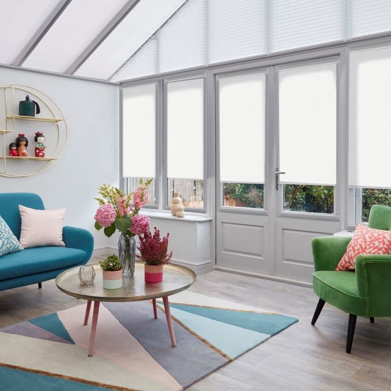 White roller blinds covering tall conservatory windows and French doors, lowered to mid-height and filtering daylight; sunroom with teal sofa, green armchair, coffee table, floral arrangement and garden view.