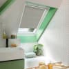 A light-gray pleated skylight blind, partially lowered across a slanted roof window, filtering daylight; attic bathroom with mint-green trim, white herringbone tiles, bathtub, sink, potted plant and toiletries.
