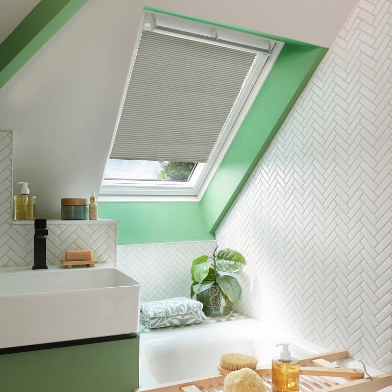 A light-gray pleated skylight blind, partially lowered across a slanted roof window, filtering daylight; attic bathroom with mint-green trim, white herringbone tiles, bathtub, sink, potted plant and toiletries.