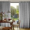Gray full-length pleated curtains with white diamond pattern, hung on a sliding glass door, drawn partly open to reveal a sunlit garden; dining area with wooden table, chair, pendant lamp.