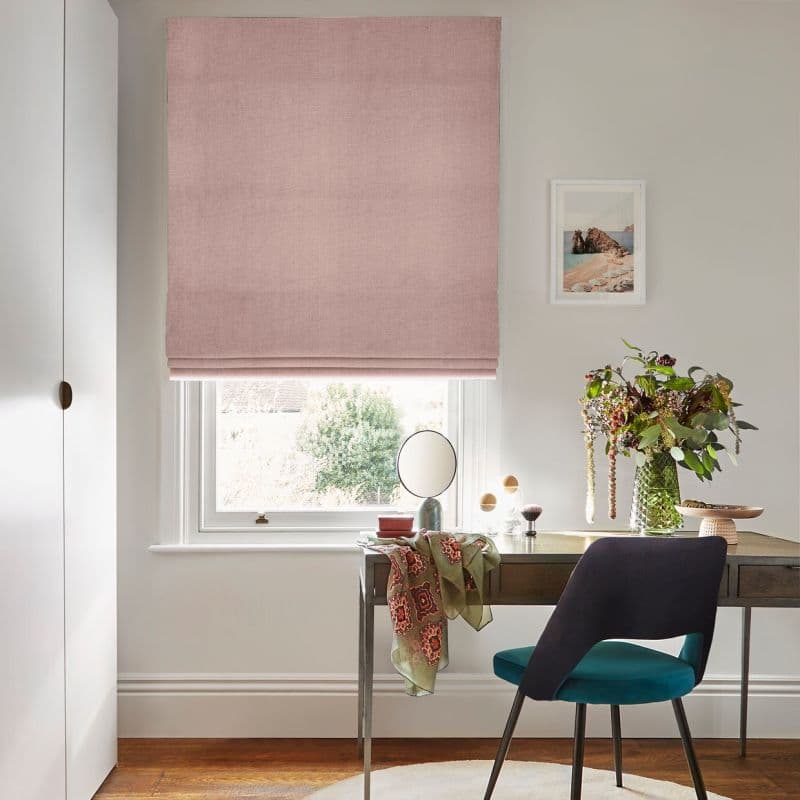 A pale pink fabric Roman blind, lowered almost fully with folds, filtering soft daylight through a bottom gap above a desk with chair and vase of flowers in an office.