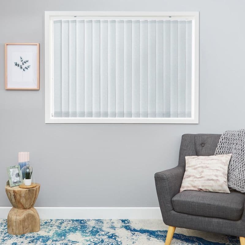 White vertical blinds cover a rectangular window, closed and diffusing light; gray living room with framed botanical print, wooden side table with decor, gray armchair, knit throw and blue rug.