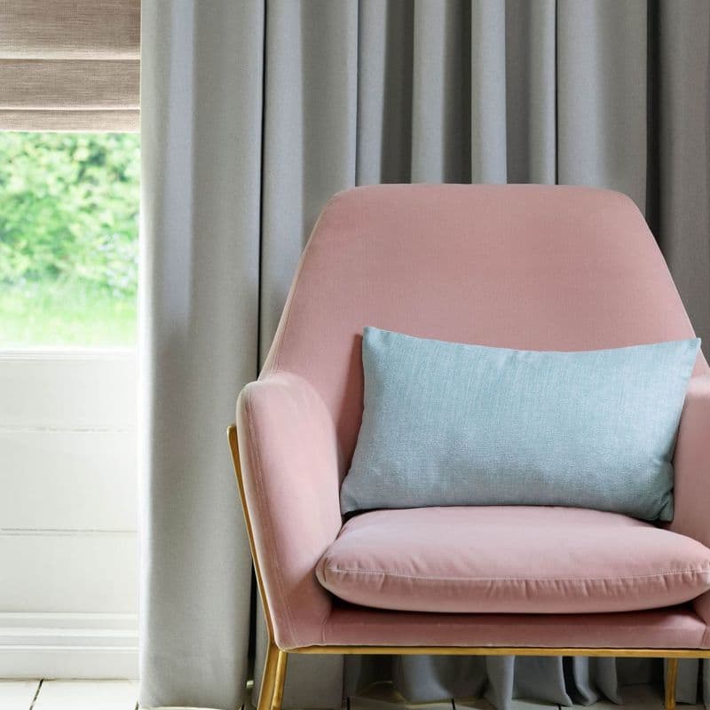Floor-length grey pleated curtains, closed beside a raised beige Roman blind filtering daylight; in a living-room corner with a pink upholstered armchair and pale blue cushion, garden visible.