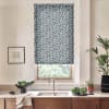 Patterned blue-and-white roller blind covers a kitchen window, filtering daylight. Below, a stainless sink and faucet sit on a wooden countertop with a towel, bowl, potted plants, and framed sketch.