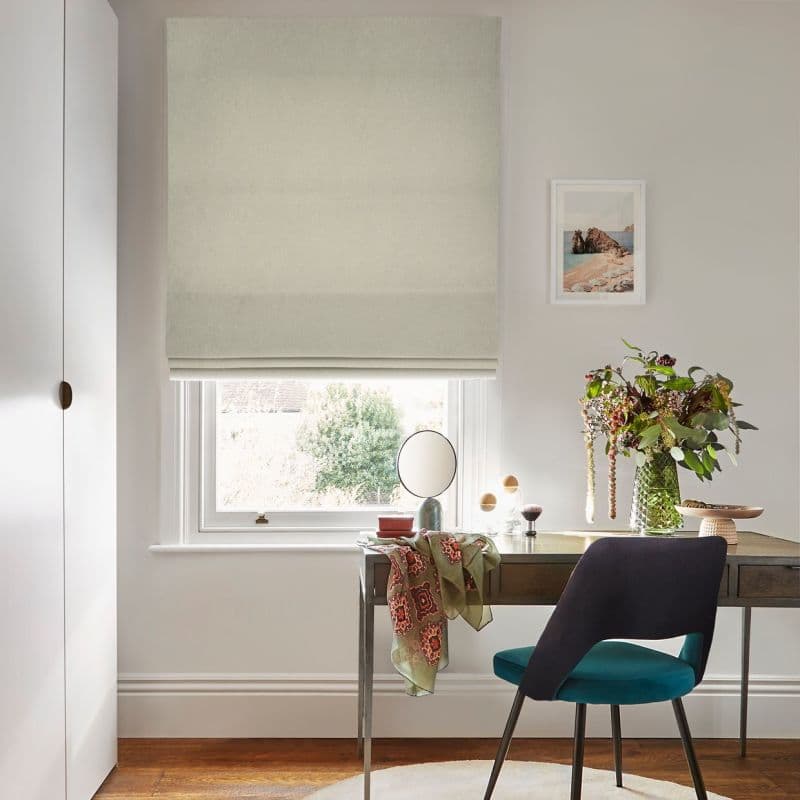 Roman fabric blind, light beige, fully lowered with soft horizontal folds, filtering daylight into a study; wooden desk, teal chair, mirror and vase of flowers beside a small framed seascape.