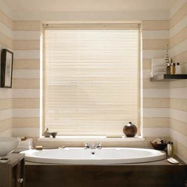 Horizontal beige Venetian blinds, fully lowered, filtering soft daylight; positioned over a window above a white oval bathtub in a softly lit bathroom with striped walls and toiletries.