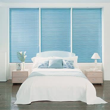 Three blue horizontal Venetian blinds covering a triple window, closed and filtering daylight; in front, a white bed with pale-blue pillows and two bedside tables in a softly lit bedroom.