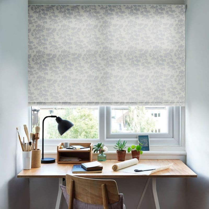 Floral-patterned roller blind, lowered fully, pale grey botanical print. Filtering soft daylight. Above a wooden desk with lamp, brushes, potted plants and notebooks; bright home-office with suburban view.