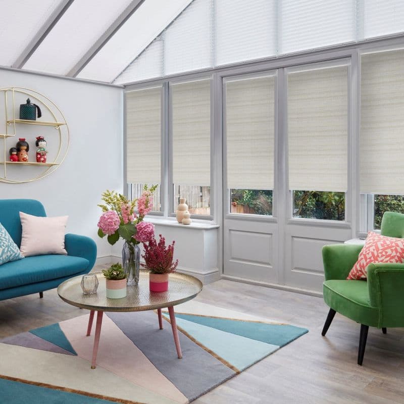 Beige fabric roller blinds lowered across tall glazed doors and upper angled windows, filtering daylight; conservatory living room with teal sofa, green armchair, round coffee table, flowers and garden view.