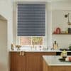 Grey textured roller blind lowered over a kitchen window, blocking light and revealing a narrow bottom gap; brass faucet and wooden cabinets on a countertop with brick wall view.