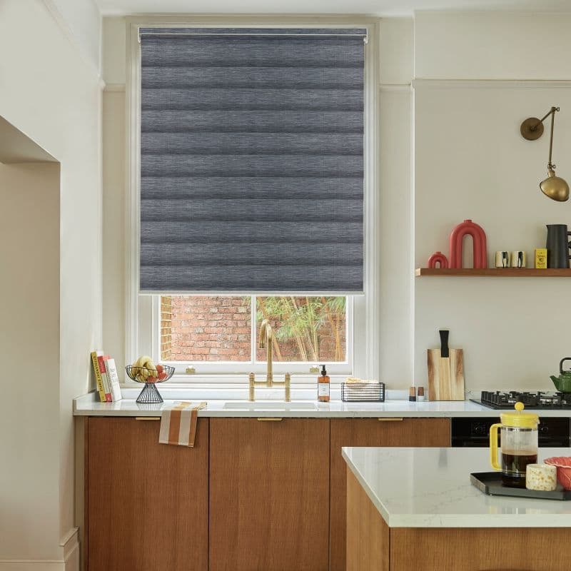Grey textured roller blind lowered over a kitchen window, blocking light and revealing a narrow bottom gap; brass faucet and wooden cabinets on a countertop with brick wall view.