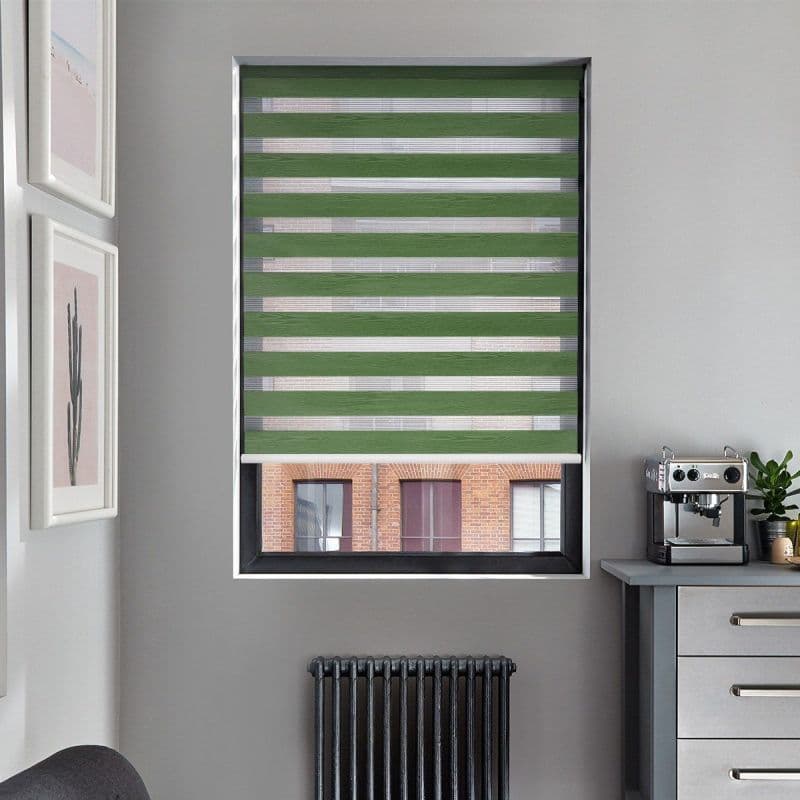 A green zebra roller blind, lowered over a window with alternating opaque stripes, filtering daylight; gray walls with radiator below, art left, espresso machine on counter right, brick building visible outside.