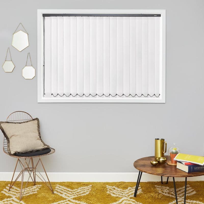 White vertical blinds inside a white-framed window with black headrail and bottom bead chain; closed, diffusing light in a grey wall living room with a wire chair and coffee table.