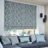 A blue-gray patterned Roman blind, lowered with a narrow bottom gap, filters daylight over a living-room window; grey sofa with cushions and pendant lights, patio garden visible beyond.