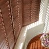Wooden plantation shutters covering a window, slats tilted, filtering sunlight and casting striped shadows across a table holding a vase of pink and white flowers in a sunlit corner.