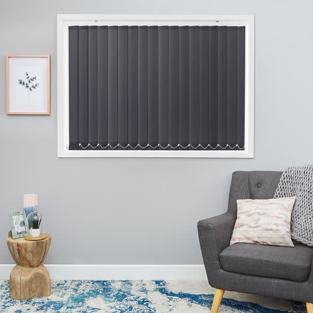 Dark grey vertical blinds fully closed, scalloped lower edges; blocking light in a grey-walled living room with a grey armchair, wooden stump side table, framed botanical print and blue rug.