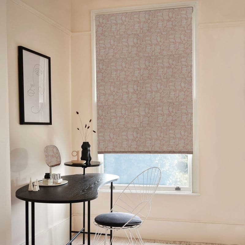 Patterned roller blind (beige with pink geometric lines) lowered, filtering light through a small lower gap; small pale study with a curved black desk, wire chair, minimalist framed line art.