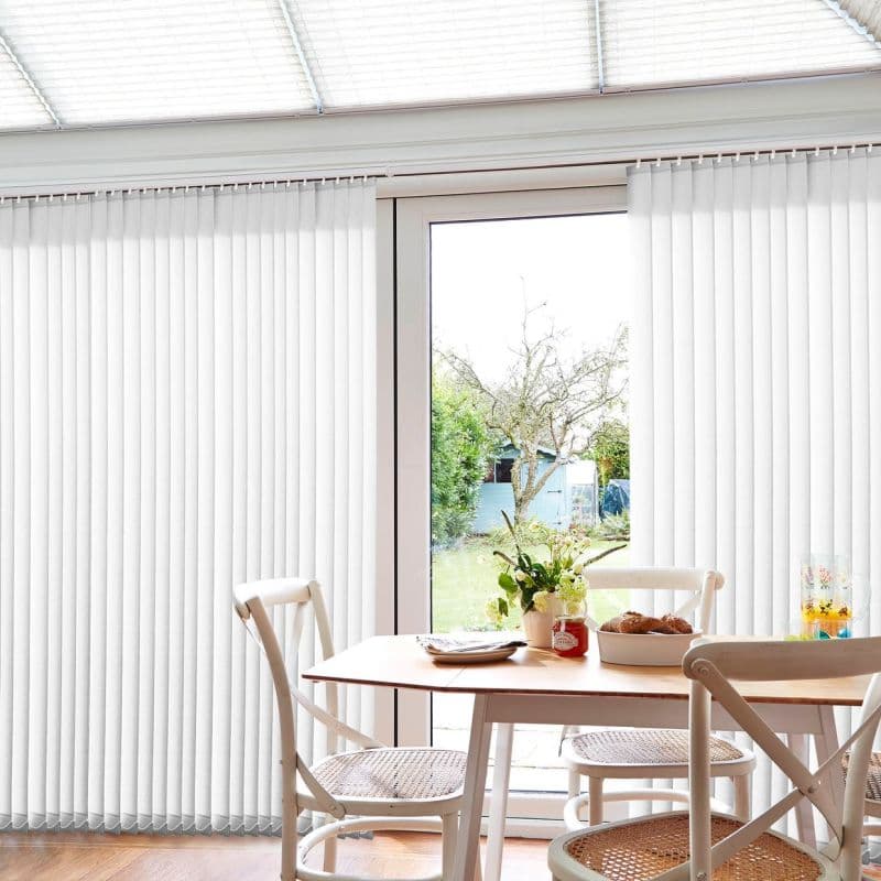 Vertical white blinds cover sliding glass doors, partially drawn at center, filtering bright daylight; wooden dining table with chairs, vase and pastries in sunlit room overlooking garden.