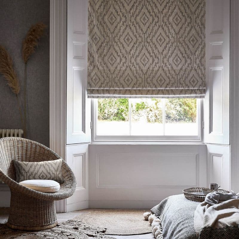 Patterned beige-grey Roman blind lowered over a window, folding at the bottom and filtering daylight; set in a white-painted window alcove of a cozy neutral sitting room with wicker chair.