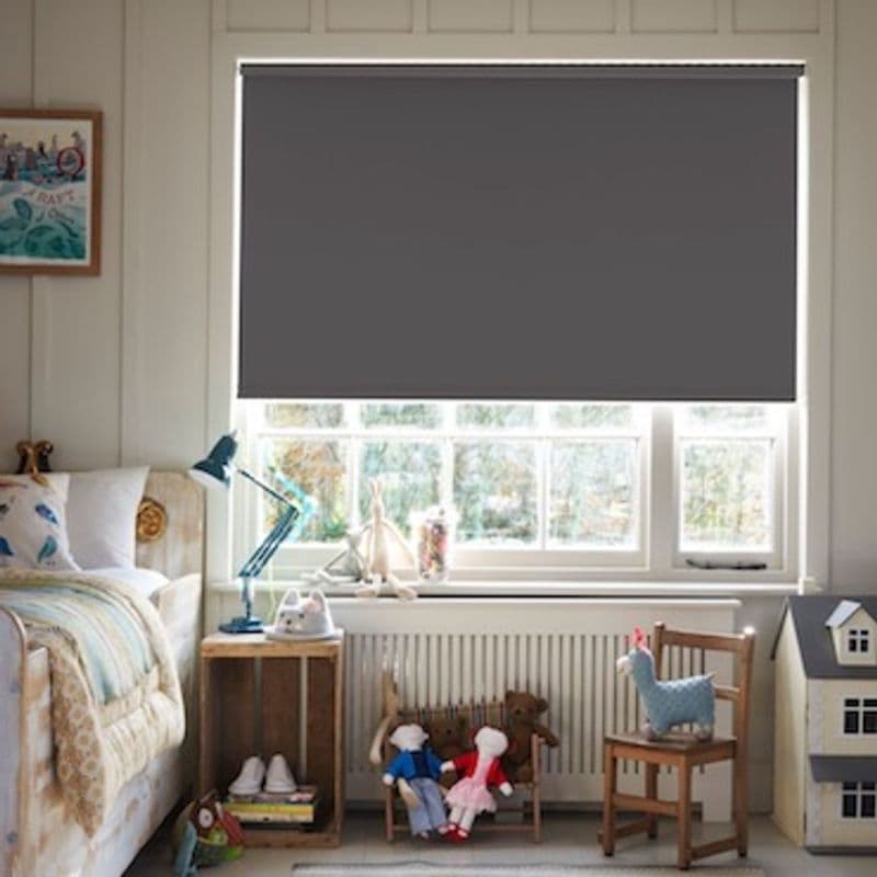 Dark gray roller blind lowered across window, edges slightly backlit. Action: blocking daylight. Context: cozy children's bedroom — bed, bedside table and lamp, dolls and toy house on chairs beneath window.