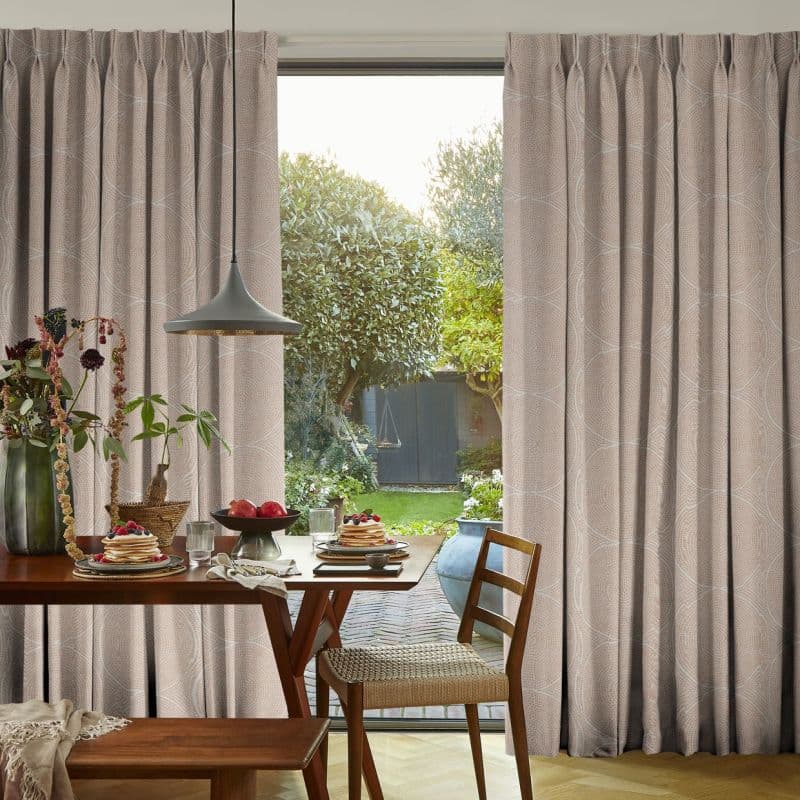 Floor-to-ceiling pleated beige curtains with subtle wave pattern, partially open to reveal a glass door; wooden dining table with plates and pendant lamp, sunlit backyard garden visible beyond.