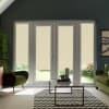 Glass patio doors with beige roller blinds lowered, framed by tall sidelights; closed handles visible. In a living room with green armchairs, glass coffee table on chevron rug, garden beyond.