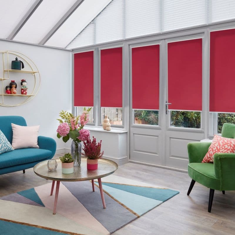 Four red roller blinds, lowered over tall glass doors and windows, covering panes; in a bright conservatory living room with teal sofa, green armchair, coffee table and pastel geometric rug.