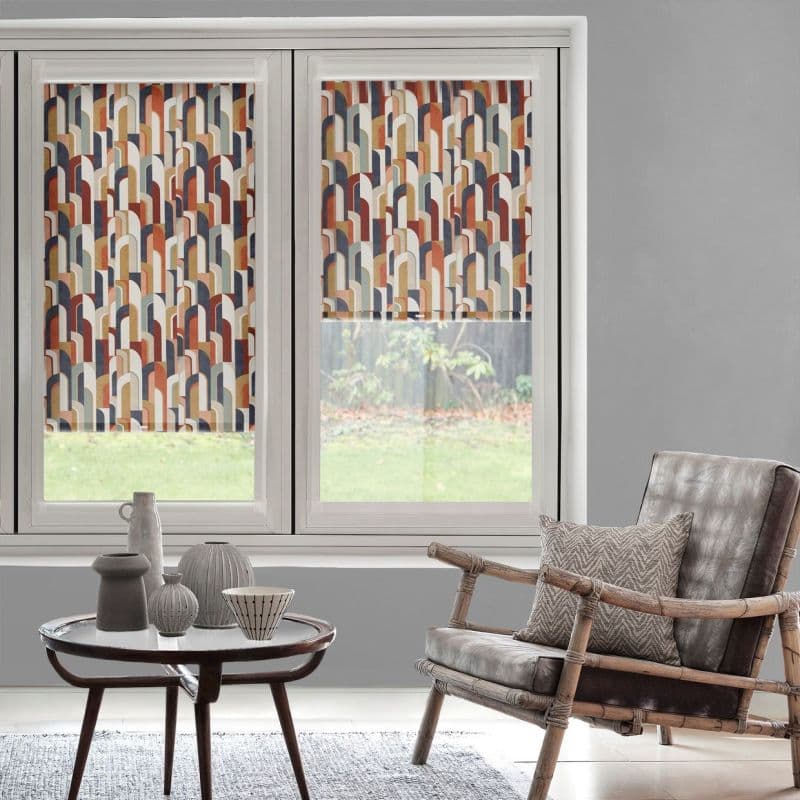 Patterned roller blinds (multicolored repeating arch motif) lowered over twin windows, filtering daylight; in a gray living room with a rattan armchair, cushion, round coffee table and garden view beyond.