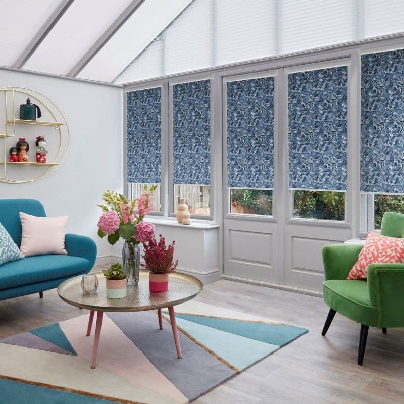 Blue swirled roller blinds, lowered to mid-height across five conservatory windows, filtering daylight; sunlit living conservatory with teal sofa, green armchair, round coffee table and floral arrangements.