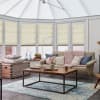 Beige pleated cellular blinds covering tall conservatory windows, lowered halfway and softly filtering daylight; a bright, cozy sunroom with a beige sofa, wooden coffee table, and blue patterned rug.