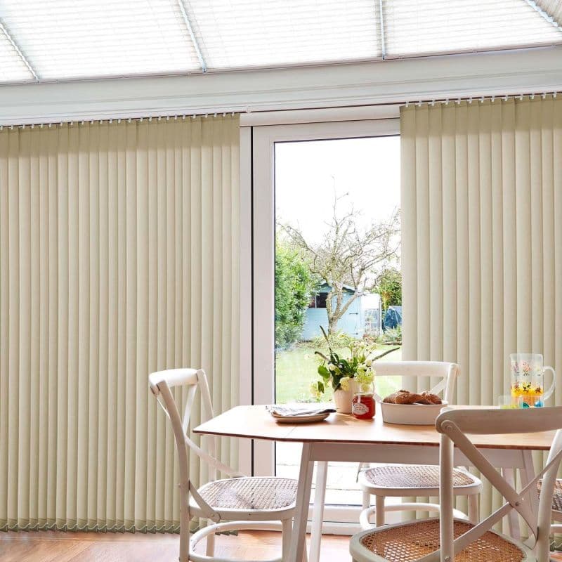 Beige vertical blinds—floor-to-ceiling slatted panels—drawn apart centrally, filtering light through a glass patio door; dining table with white chairs in a bright room, backyard with tree and shed visible.