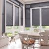 Gray horizontal Venetian blinds covering tall bay windows and French doors, partially lowered and closed, filtering daylight into a bright dining area with wooden table, chairs, and garden view.
