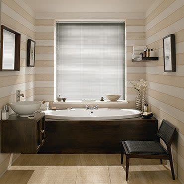 Horizontal venetian blinds covering a central bathroom window, fully lowered and closed, softly diffusing daylight; modern tub beneath, wooden vanity, striped pale walls, small shelf, chair and decorative vase.